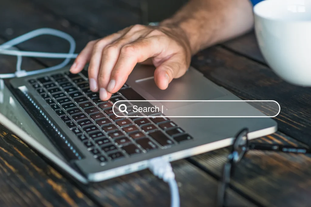 Mão usando notebook com busca na internet, digitando no teclado e conectando um cabo, em uma mesa de trabalho, representação de site no google