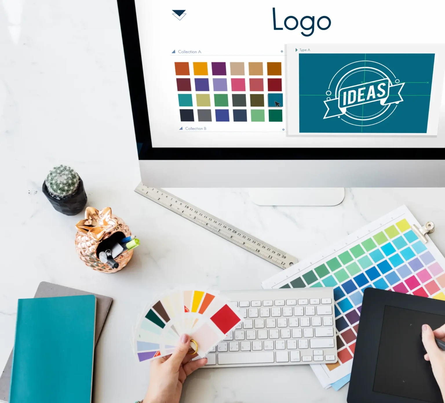 Mesa de trabalho de design com computador exibindo tela de “Logo”, paleta de cores, formas e amostras de identidade visual, além de teclado, tablet e anotações.